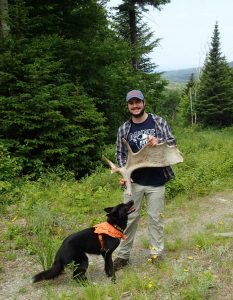 2019 Swan Intern in the woods holding a moose shed he found with a black dog standing next to him.